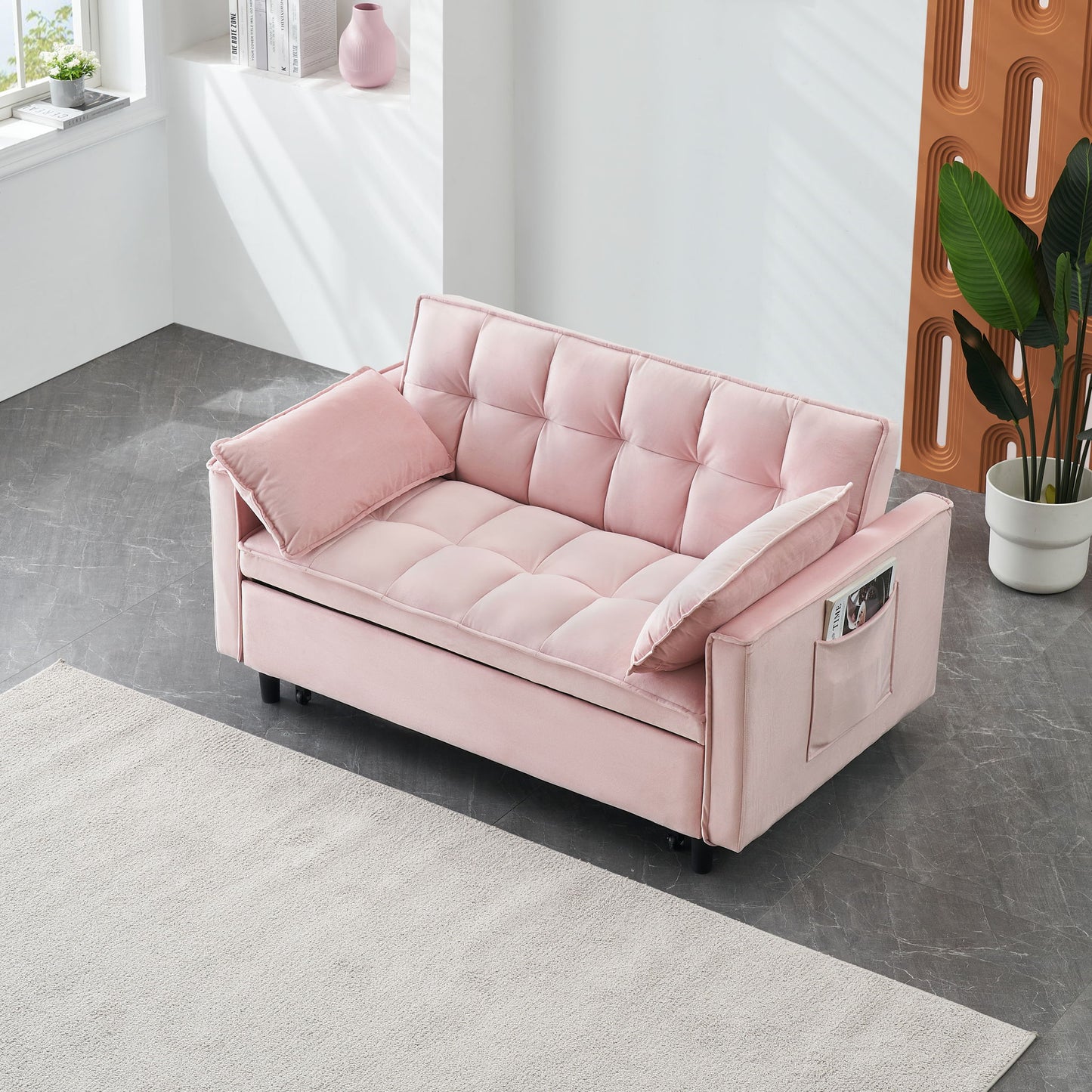 A modern room with the Modern Velvet Sofa 3-in-1 convertible sleep sofa bed in light pink, featuring side pockets and pillow, placed on a gray floor near a window, white wall with vase, large green plant, and beige rug.
