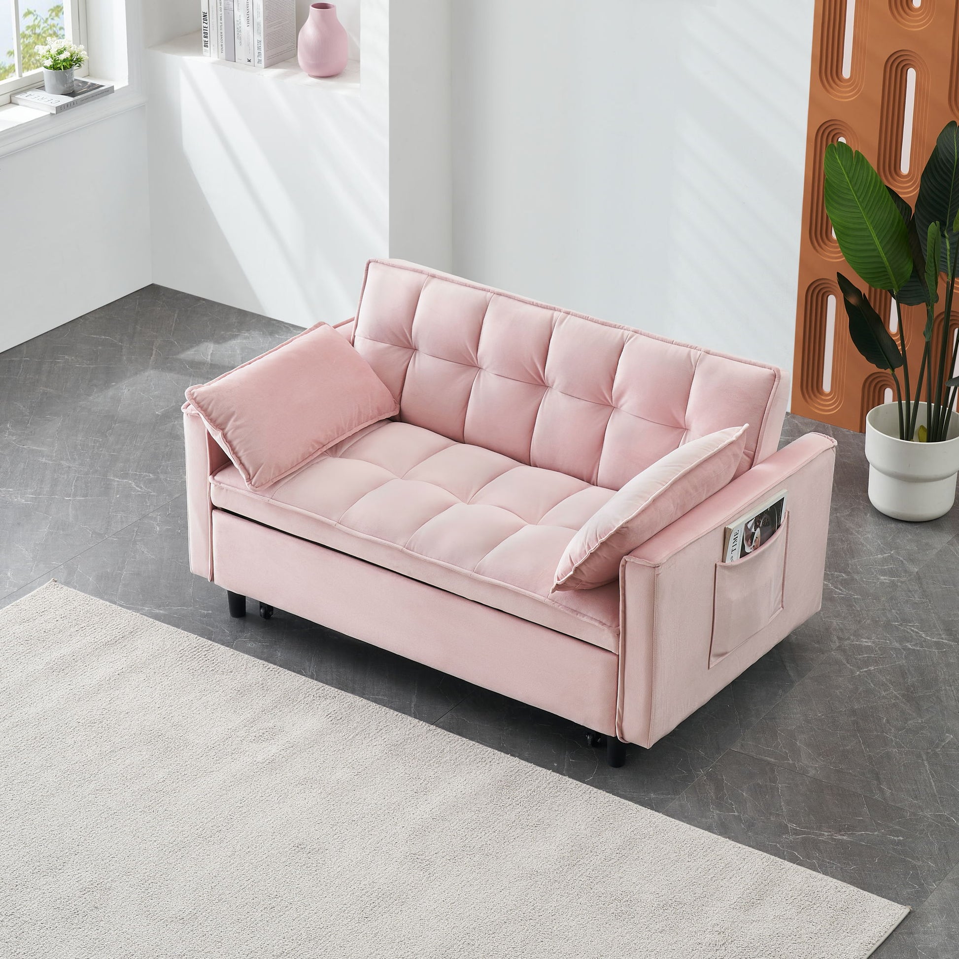 A modern room with the Modern Velvet Sofa 3-in-1 convertible sleep sofa bed in light pink, featuring side pockets and pillow, placed on a gray floor near a window, white wall with vase, large green plant, and beige rug.