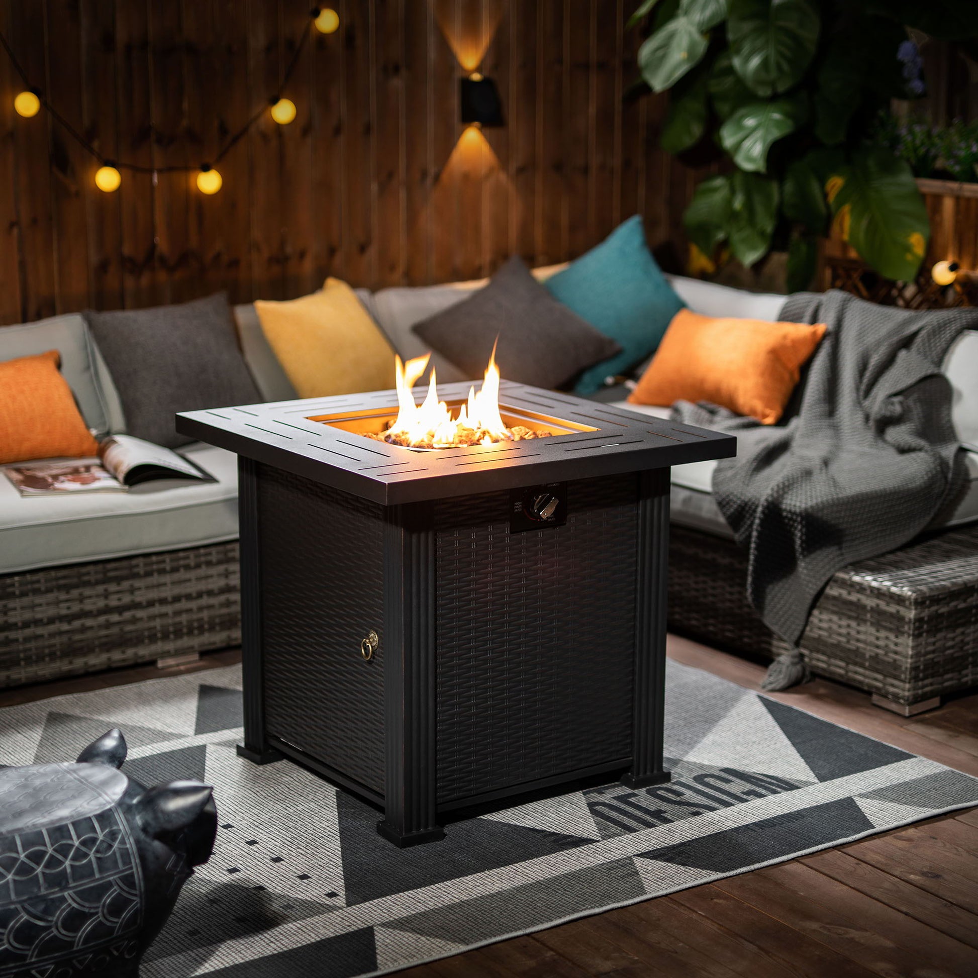 A cozy patio features the 28" Square Fire Pit Table - Brown, glowing at the center of a sectional sofa with colorful cushions and a gray blanket. String lights and plants add warmth to the wooden-fenced outdoor space.