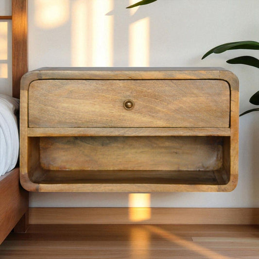 The Curve Upper Slot Floating Nightstand, a modern wooden wall-mounted piece with a drawer and lower open shelf, sits beside a bed. Sunlight creates soft shadows, while green plant leaves are visible to the right.