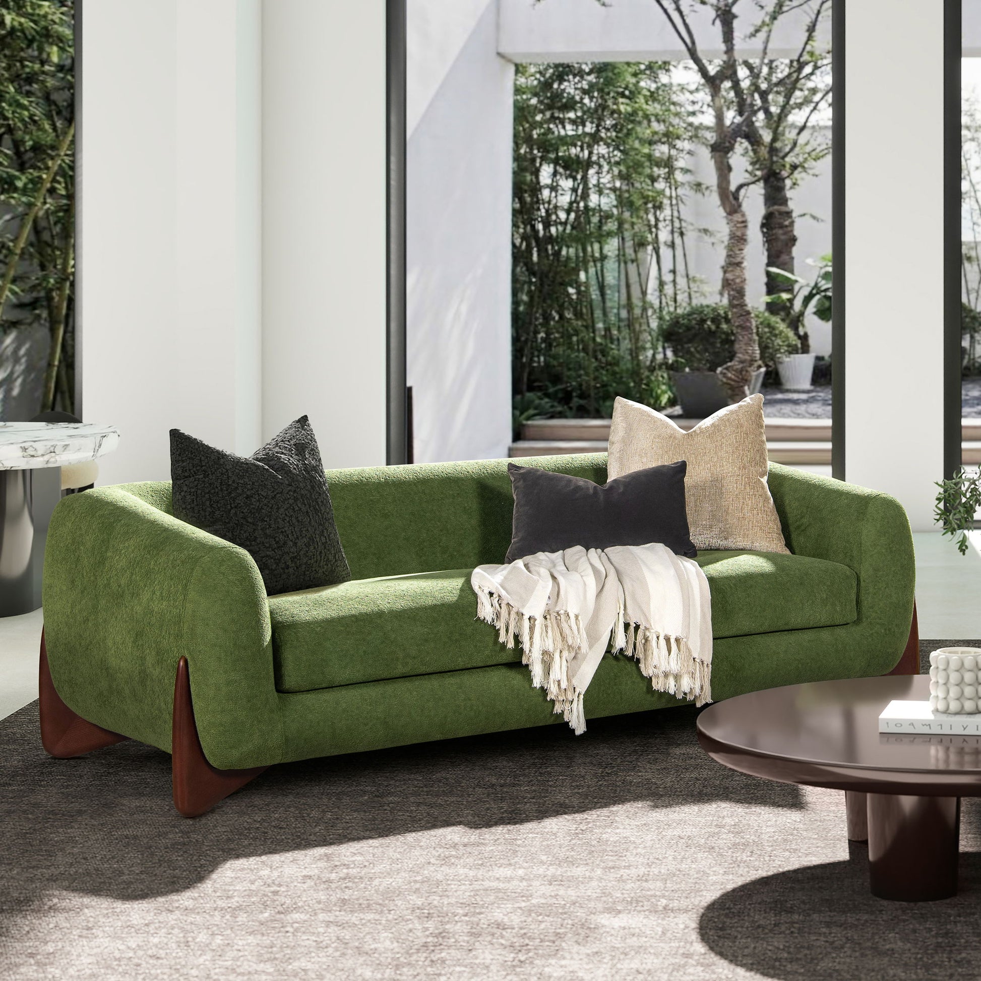 A modern living room with the Alpine - Luxurious Upholstery Sherpa Sofa in green, dark wooden legs, three throw pillows, a white fringed blanket, set on a gray rug near a round brown coffee table and large windows overlooking a garden.
