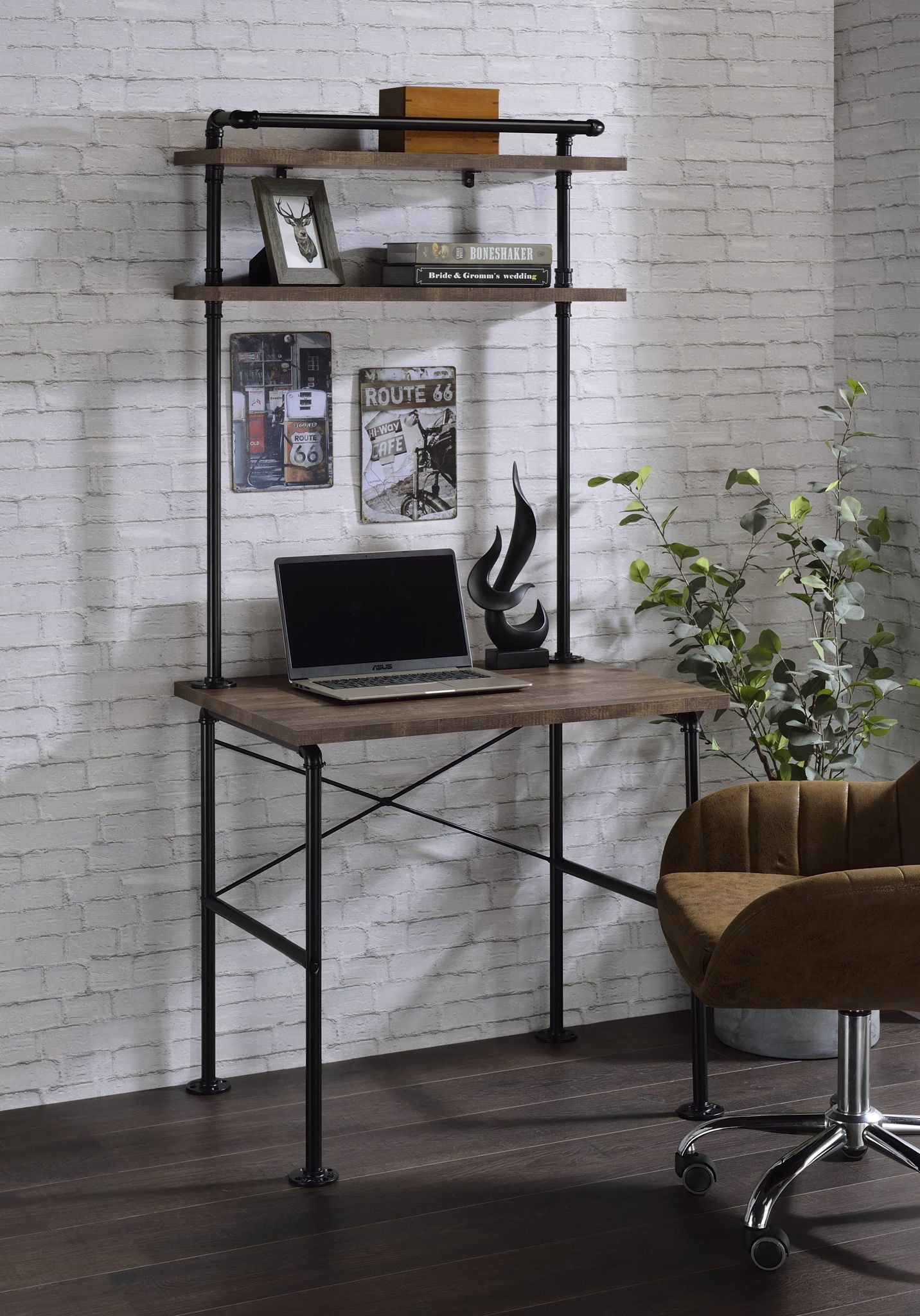 A home office setup featuring the Ensata Rustic Writing Desk in black/oak, shelves, a laptop, modern black sculpture, books, framed photo, Route 66 sign, small decor items, potted plant, and a brown swivel chair against a white brick wall.