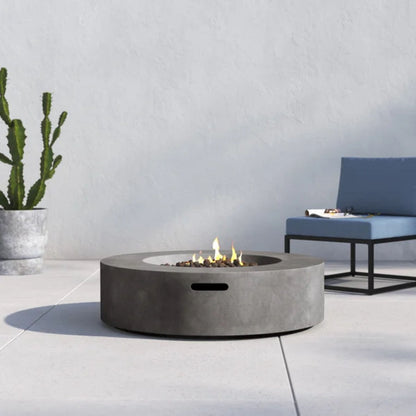 A modern outdoor scene featuring the Fiber Reinforced Outdoor Fire Pit Table in black/gray, a potted cactus, and a blue cushioned chair with black metal frame on a light-colored patio.
