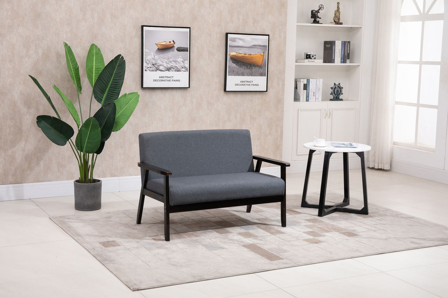 A modern living room featuring the Bahamas - Loveseat in gray, a round white side table, potted green plant, two framed boat photos on the wall, beige rug, and white shelves displaying decor and books.