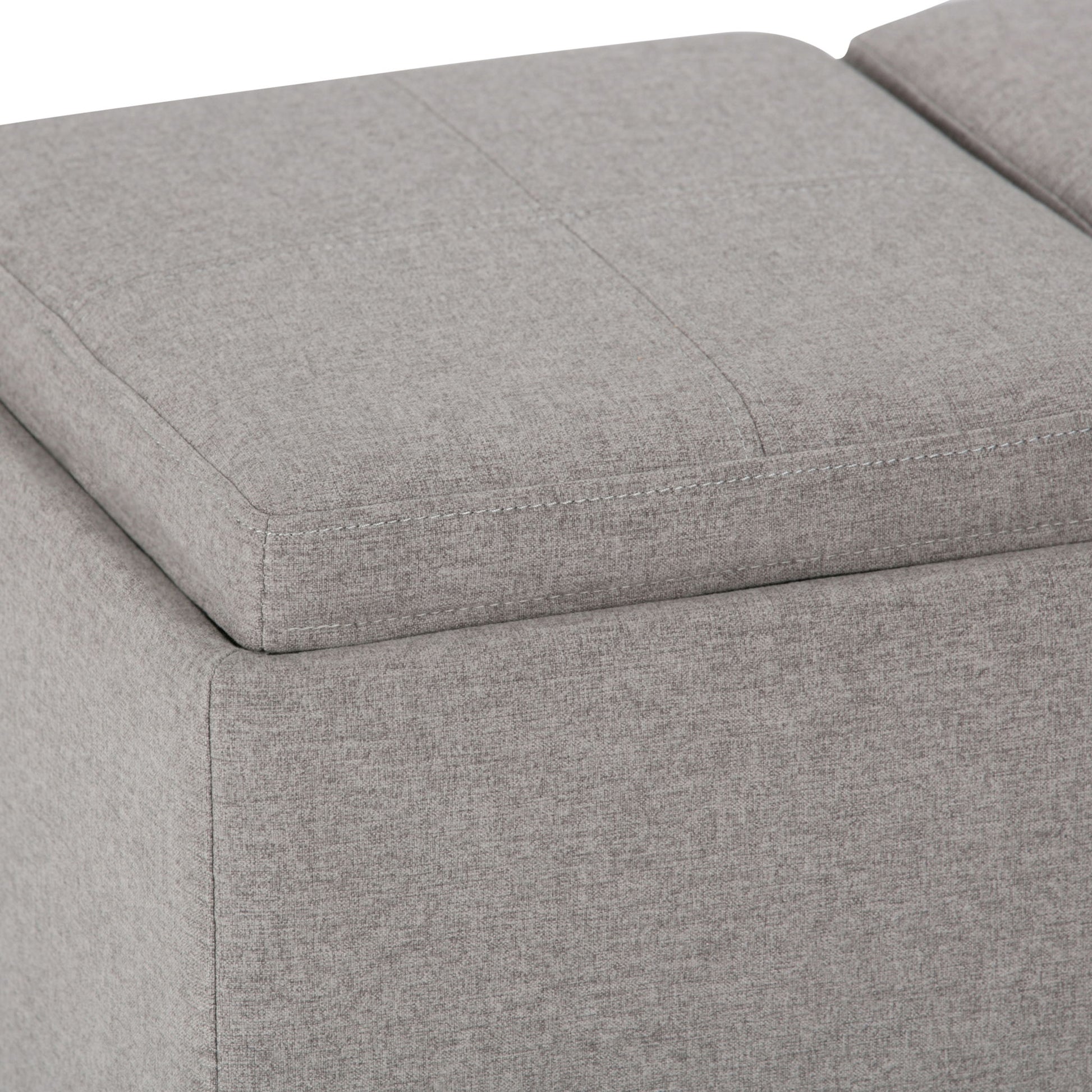 Close-up view of the Avalon Upholstered Storage Ottoman in light gray, featuring visible stitching and a removable cushioned top.