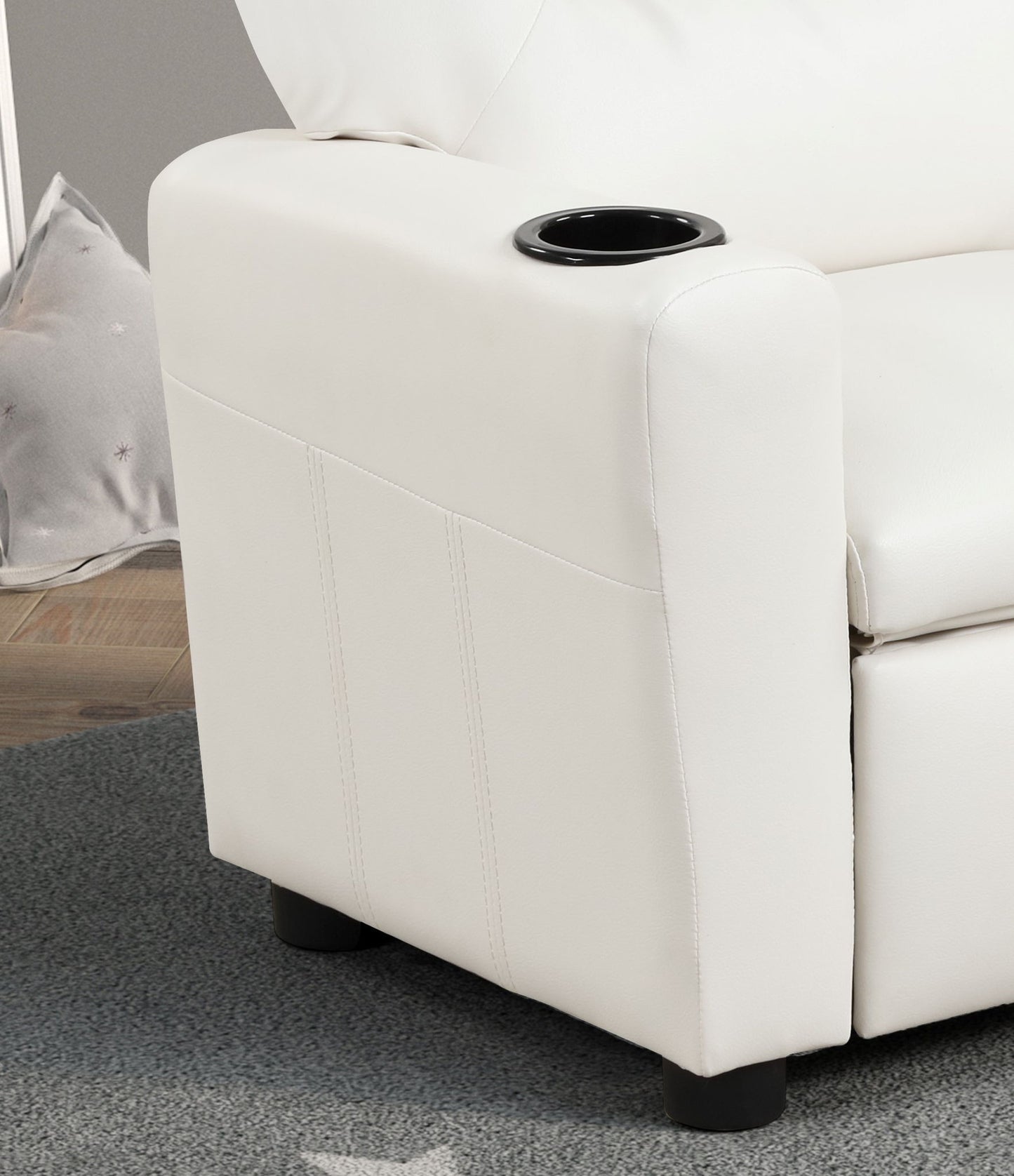 A close-up of the Marisa - PU Leather Kids Recliner Chair With Cupholder shows its modern white armrest with a built-in black cup holder, set on a gray carpeted floor. A pillow is partially visible in the background.