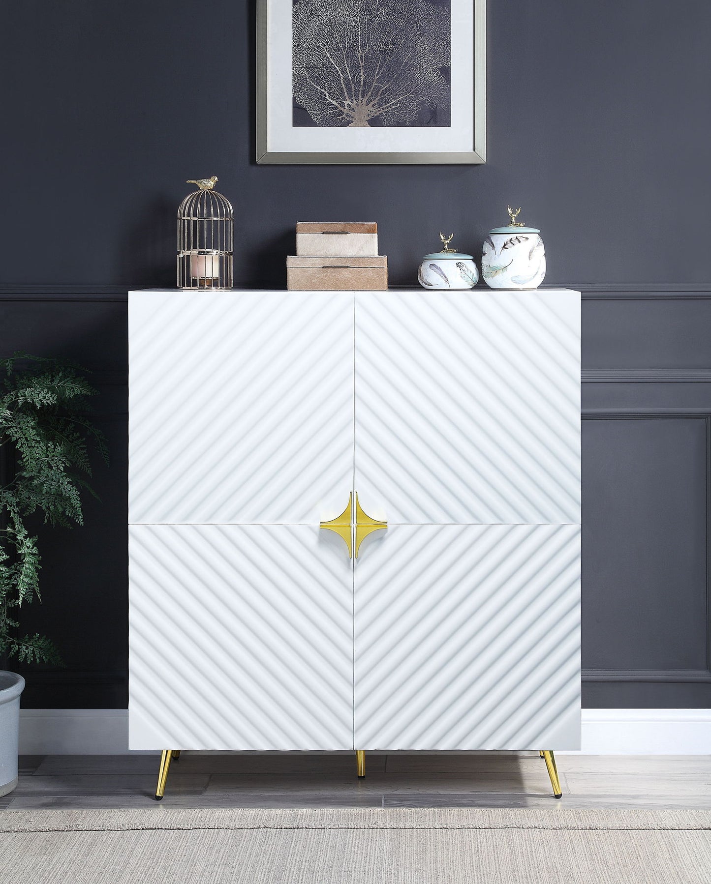 The Gaines High Gloss Accent Cabinet features modern white diagonal-textured doors, gold handles, and legs. It stands against a dark wall, topped with decor like a birdcage, boxes, jars, and is accented by framed artwork above.