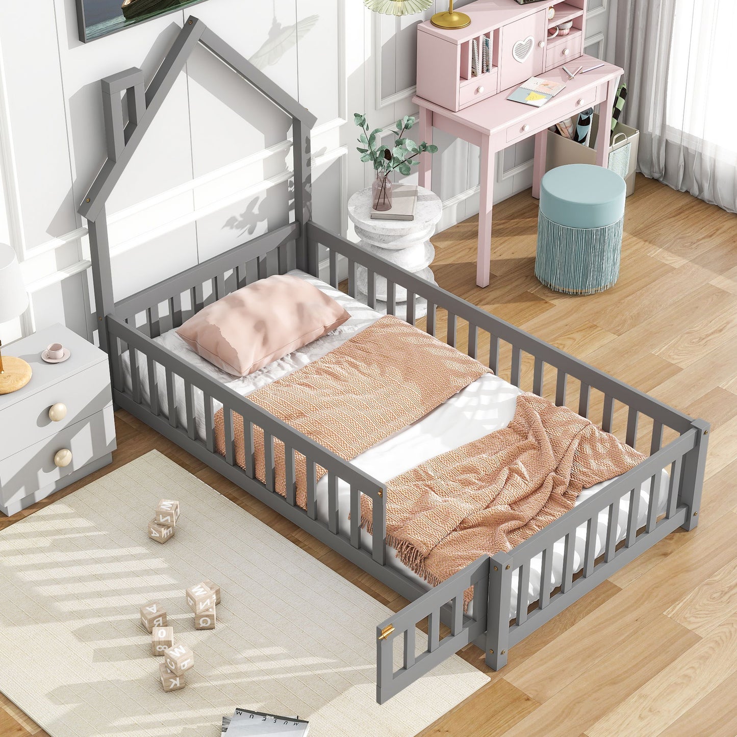 A child's bedroom featuring the House-Shaped Headboard Floor Bed With Fence in gray, pink bedding, a matching desk with shelves, blue ottoman, white rug on wooden floors, and sunlight through sheer curtains.