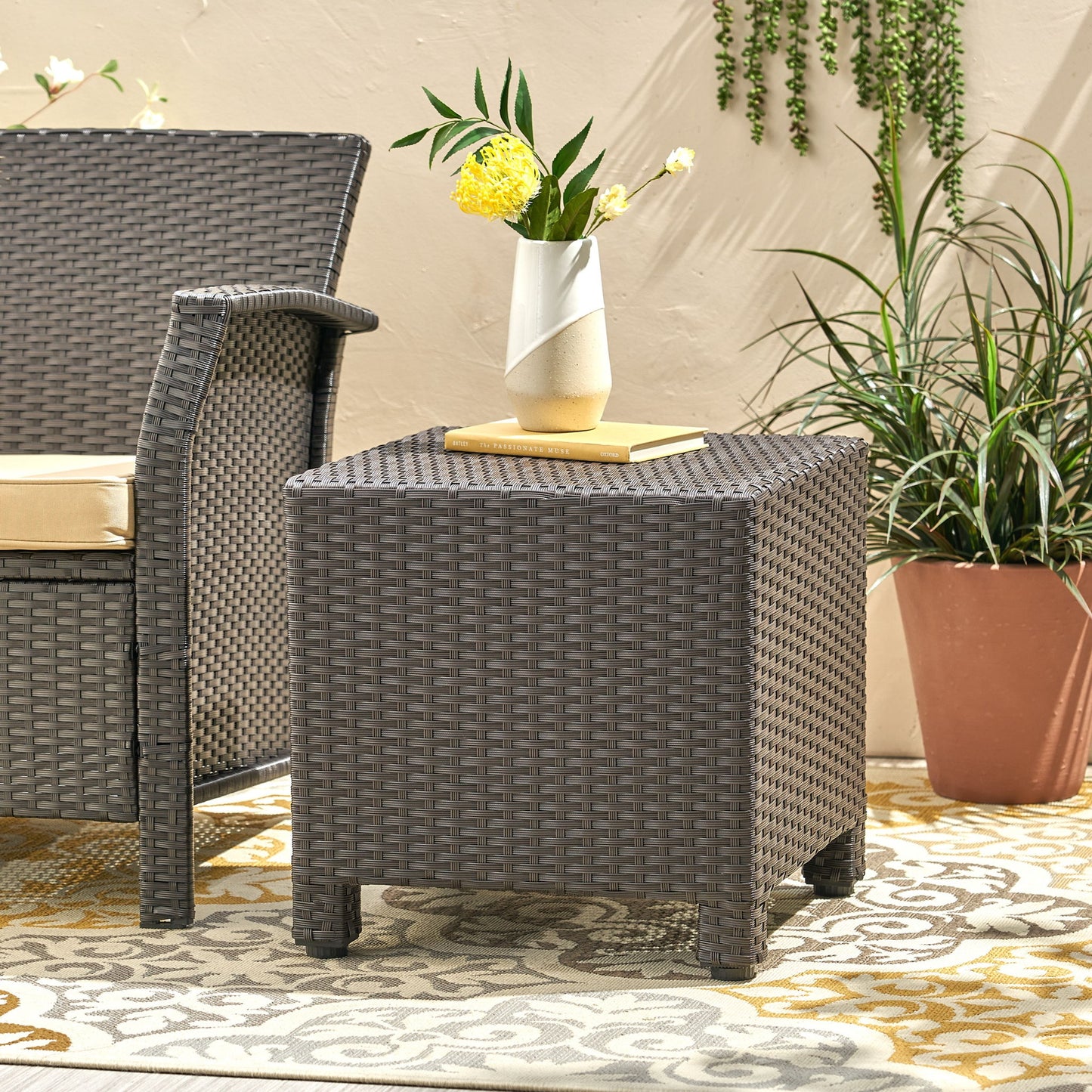 The Puerta - Minimalist Style Accent Table holds a vase of yellow flowers and a book beside a matching cushioned chair, set on a patterned rug with potted plants in the background.