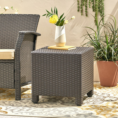 The Puerta - Minimalist Style Accent Table holds a vase of yellow flowers and a book beside a matching cushioned chair, set on a patterned rug with potted plants in the background.