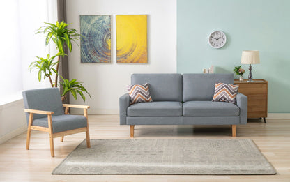 A modern living room featuring the Bahamas Sofa Set with 2 throw pillows, a matching armchair, a wooden side table with a lamp, a green plant, wall clock, two abstract paintings, and a patterned rug on light wood flooring.