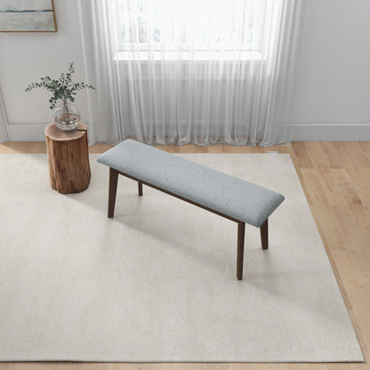 A minimalist room features a light rug, wooden floor, the Carlos Upholstered Bench in gray, a small wooden side table with a glass vase and greenery, and sheer white curtains over the window.