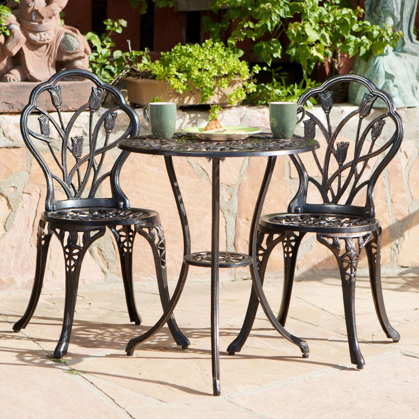 The Nassau - Cast 3 Piece Bistro Set, featuring two chairs and a round table in black metal, is shown on a stone patio with green mugs and potted plants, set against lush greenery and a stone wall in the background.
