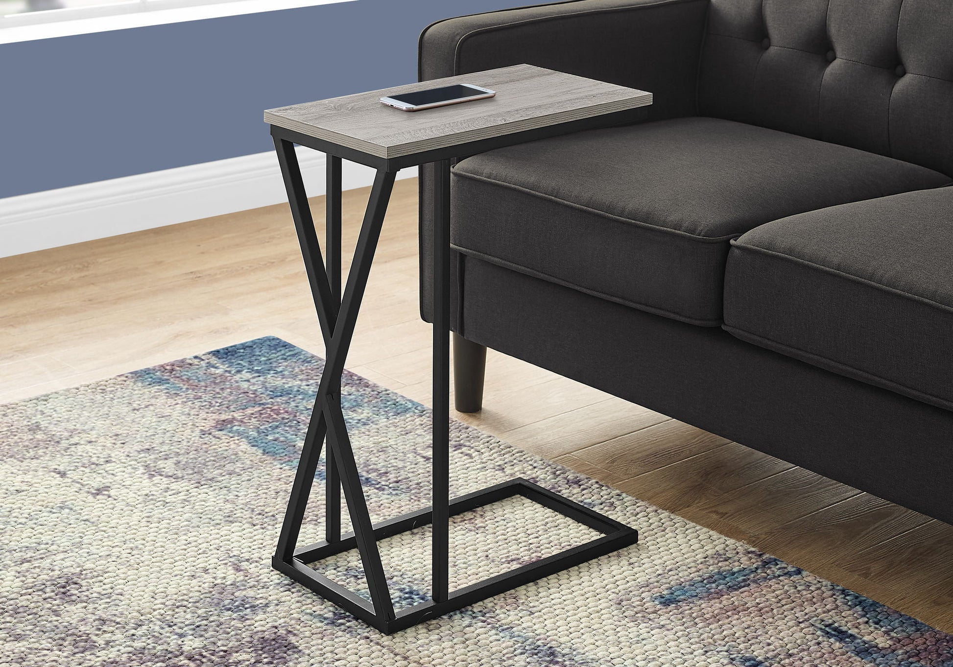 The Accent Table C-Shaped, Contemporary & Modern with a black metal frame and rectangular wooden top sits by a dark gray sofa, holding a smartphone. A colorful rug is partly visible on the wooden floor nearby.