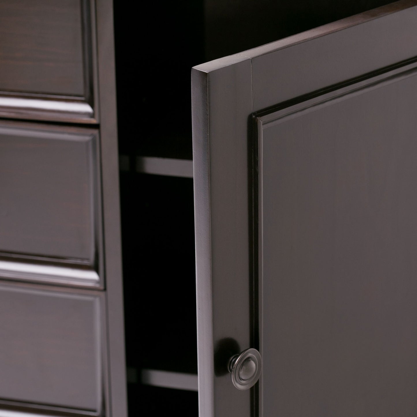 Close-up of the Amherst - Handcrafted Medium Storage Cabinet in dark wood, featuring a partially open door with a round knob revealing shelves inside, and three panel drawers visible on the left.