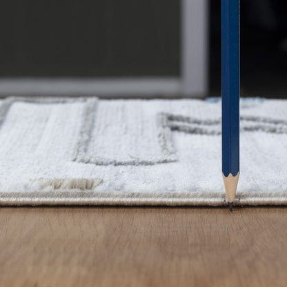 A blue pencil stands upright on a wooden floor, its tip touching the ground near the edge of the Legacy - GC_CAM8001 Area Rug, which features a light color and textured pattern.
