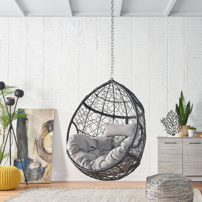 A modern living room features the Teardrop Hanging Chair With 8Ft Chain with gray cushions, a woven pouf, indoor plants, abstract art, a floor lamp, and light wood furniture set against a white brick wall.