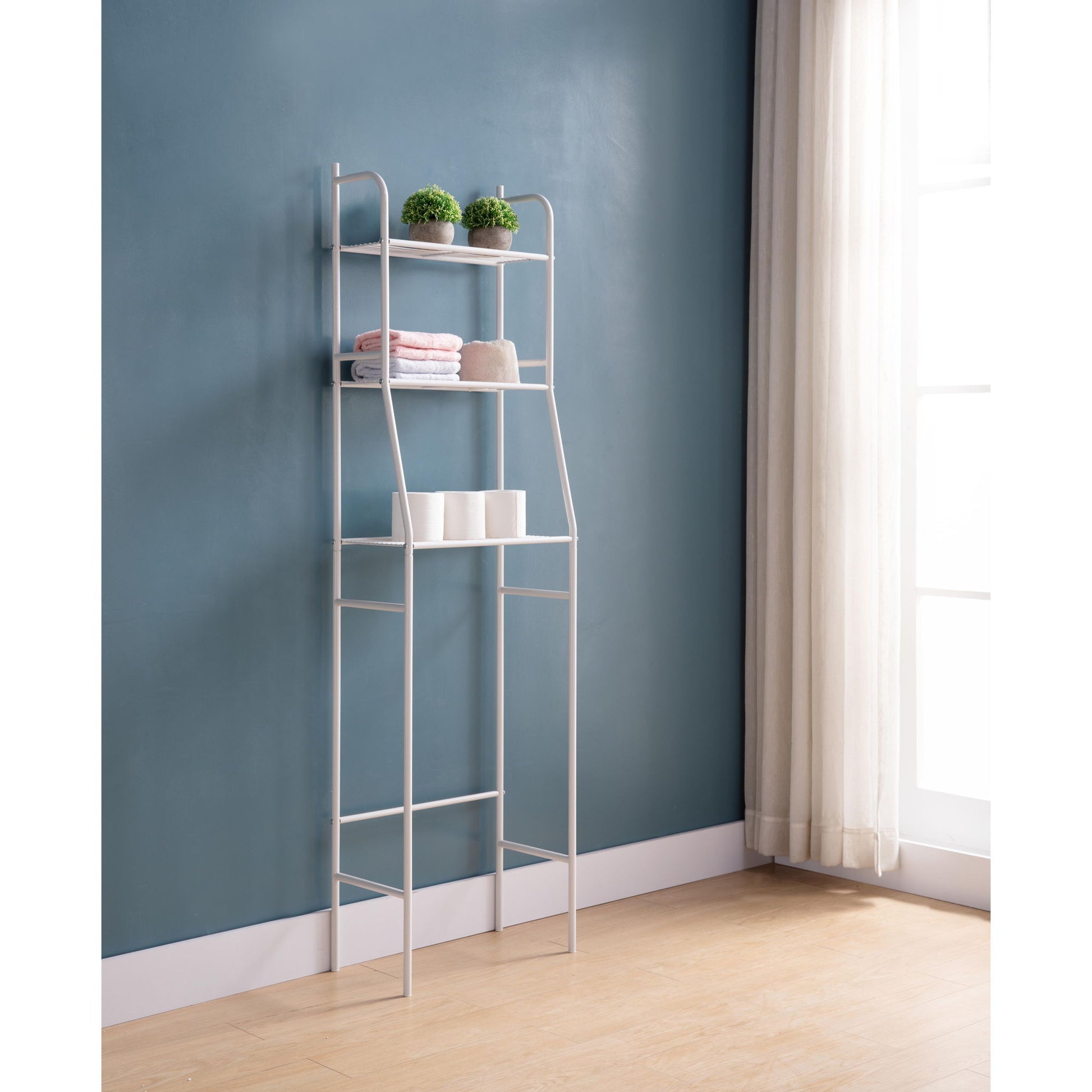 A Three Tier Over The Toilet Storage Shelf - Wood stands against a blue wall by a window with white curtains, displaying potted plants, folded towels, and three white cups on a wooden floor.
