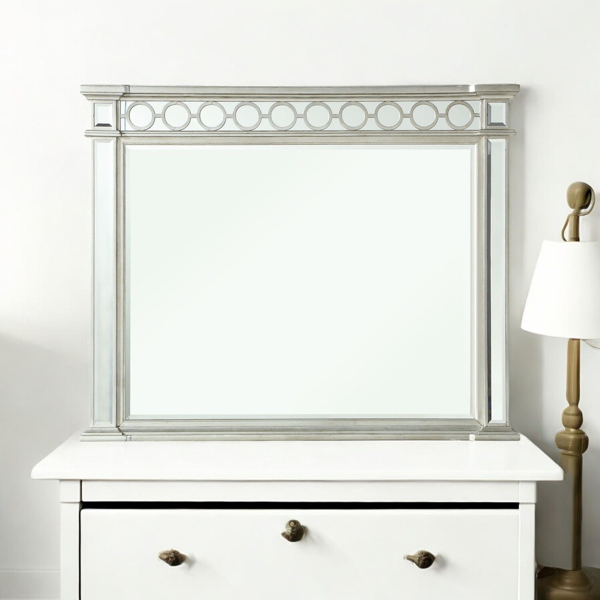 The Varian - Mirrored Designed Mirror in silver, featuring a circular top pattern, sits on a white dresser beside a brass and white lamp against a plain white wall.