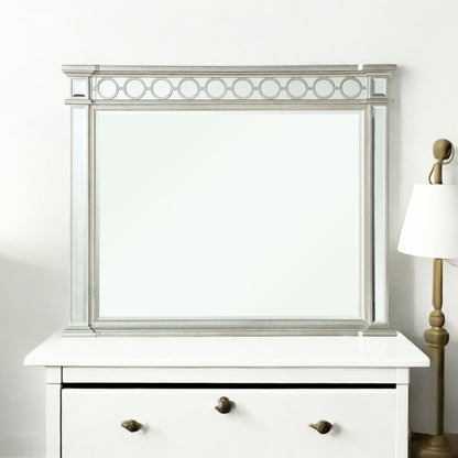 The Varian - Mirrored Designed Mirror in silver, featuring a circular top pattern, sits on a white dresser beside a brass and white lamp against a plain white wall.