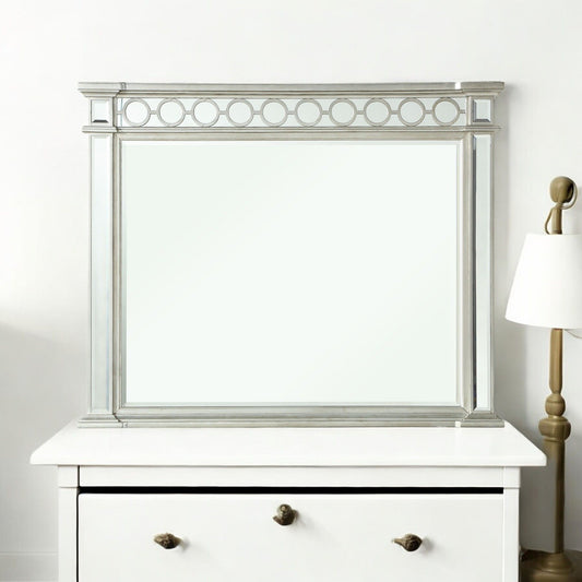 The Varian - Mirrored Designed Mirror in silver, featuring a circular top pattern, sits on a white dresser beside a brass and white lamp against a plain white wall.