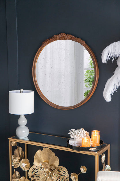 The Round Hand Carved Rose Antique Mirror Frame, Wall Mirror For Live Space, hangs on a dark wall above a gold-accented cabinet topped with a white lamp, coral decor, candles, and a tray. White curtains and greenery reflect in the mirror.