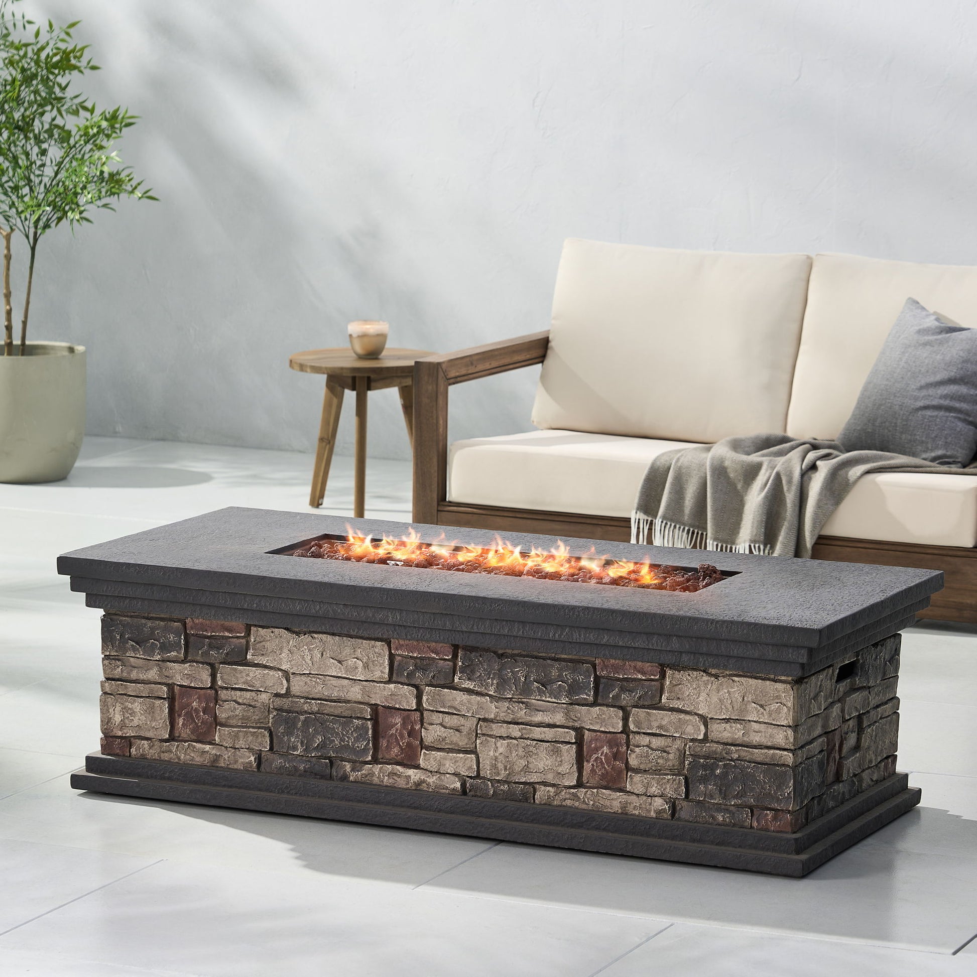 A Fire Pit With Lightweight Concrete And Steel Frame burns in front of a wooden outdoor sofa with cream cushions and a gray blanket, set on a patio featuring a potted plant and a light gray wall in the background.