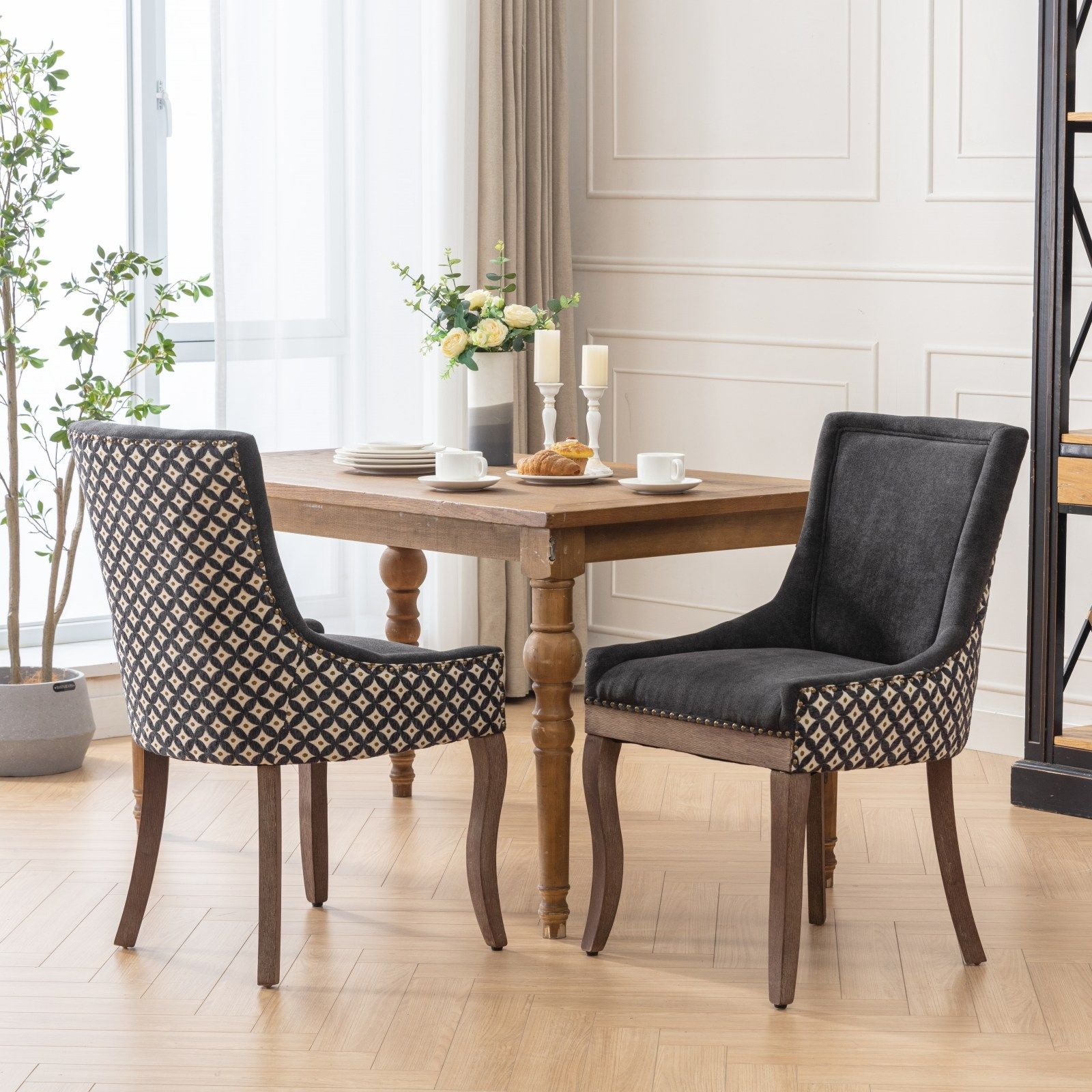 A bright, elegant room features a wooden dining table with dishes and pastries, flanked by two Ultra Side Dining Chairs—thickened fabric seats, neutral wood legs, bronze nail heads—plus large windows, plants, and a flower vase on the table.