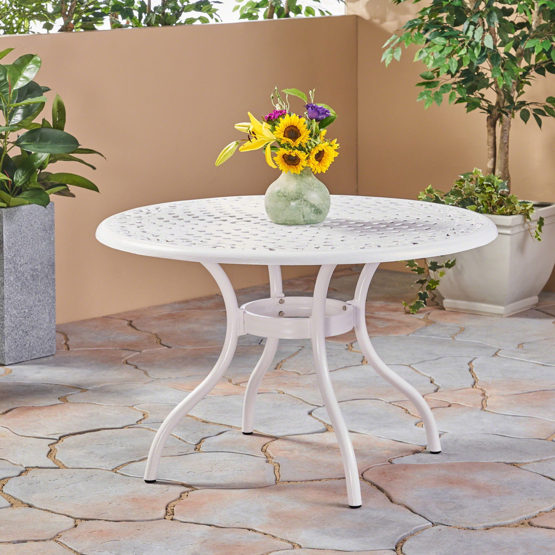 The Tranquilius Outdoor Round Dining Table with a lattice tabletop, umbrella hole, and curved legs is set on a stone patio, topped with a green vase of sunflowers and purple flowers, and surrounded by potted plants.