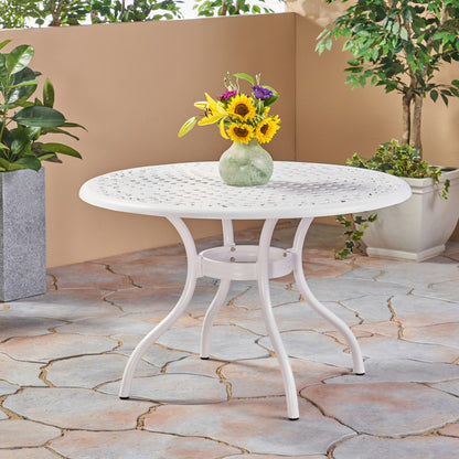 The Tranquilius Outdoor Round Dining Table with a lattice tabletop, umbrella hole, and curved legs is set on a stone patio, topped with a green vase of sunflowers and purple flowers, and surrounded by potted plants.