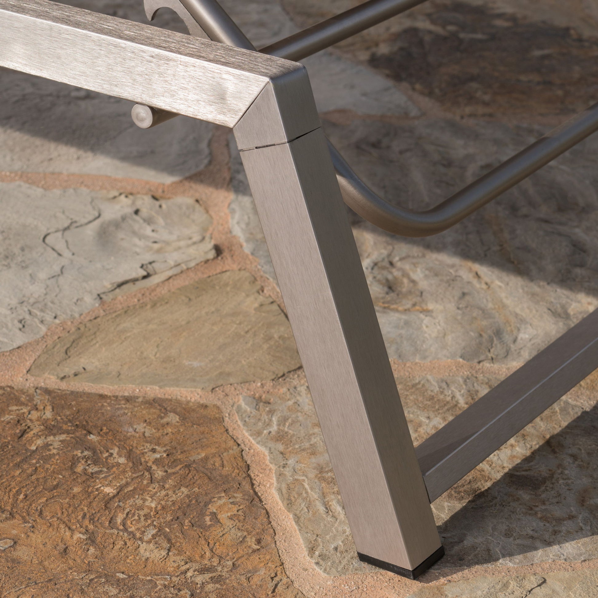 Close-up of the Myers - Modern Chaise Lounge Set With Adjustable Mesh Back, showing its brushed metal leg resting on textured, earthy-toned stone tiles.