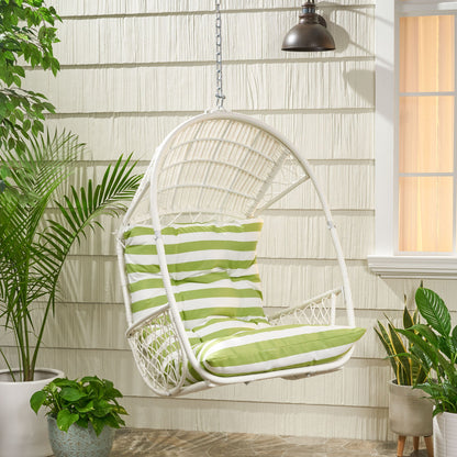 The Malia - Wicker Hanging Chair With Cushion, featuring green and white striped cushions, hangs by a chain on a covered patio with potted plants and is illuminated by a black outdoor lamp near the window.