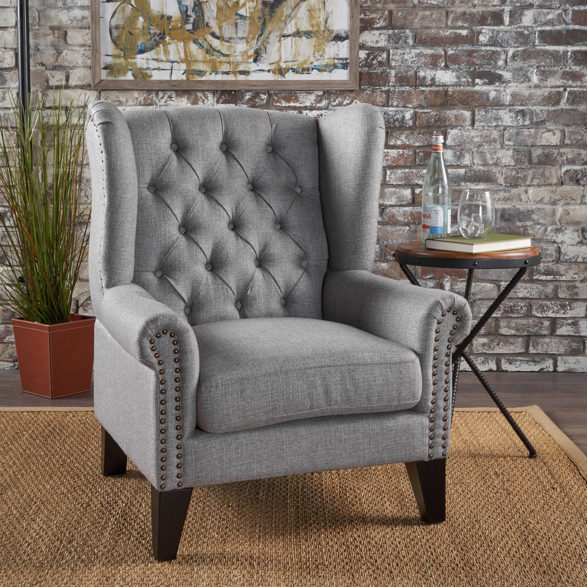 A Mid Century Modern Arm Reading Chair sits on a woven rug. Next to it is a round side table with a water bottle, two glasses, and books. A potted plant and brick wall serve as the background.