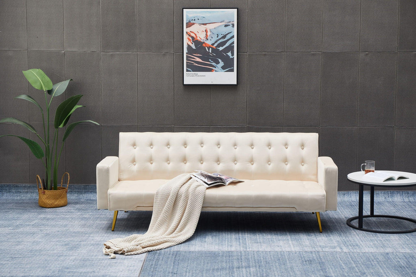 A modern living room featuring the Comfort Compact Design Sofa Bed in cream, a beige knit blanket, open magazine, round white side table with book and glass, potted plant, and framed abstract artwork on a dark wall.