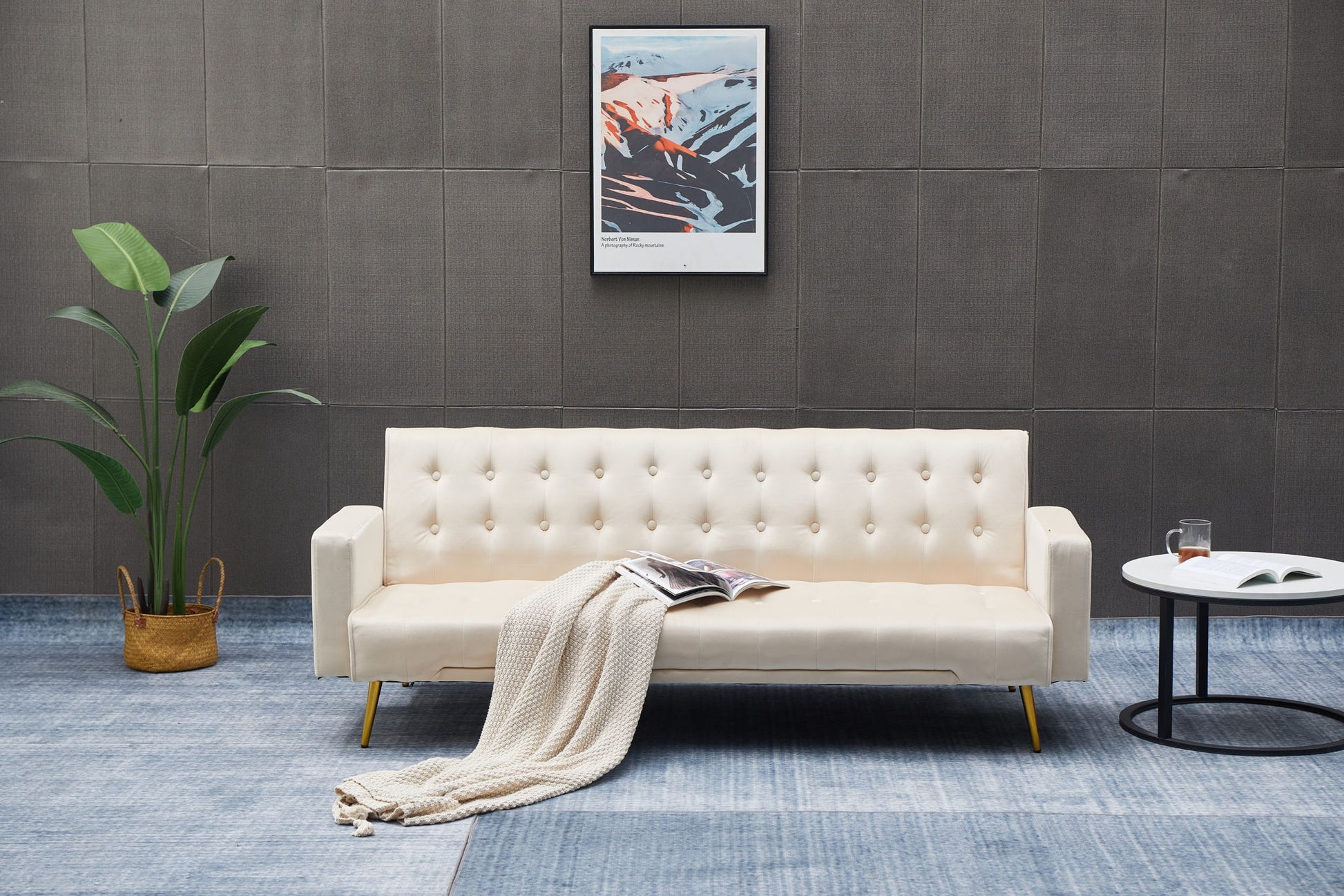 A modern living room featuring the Comfort Compact Design Sofa Bed in cream, a beige knit blanket, open magazine, round white side table with book and glass, potted plant, and framed abstract artwork on a dark wall.