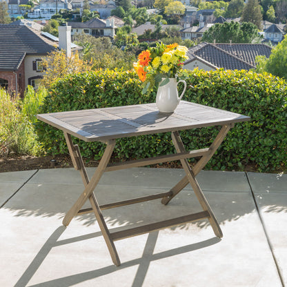 The Blissique Outdoor Foldable Wood Dining Table with a slat design sits on a patio, holding a pitcher of yellow and orange flowers, backed by green shrubs and facing a suburban neighborhood.