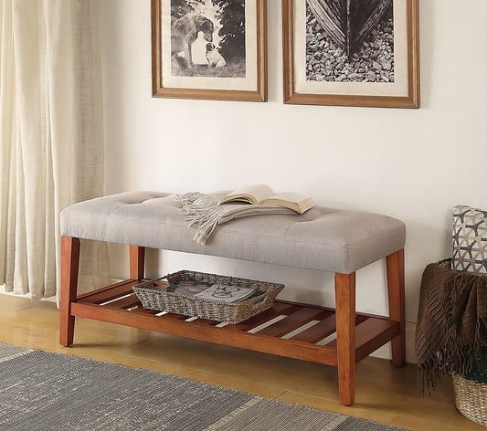 The Charla Upholstered Bench with a cushioned beige seat sits against a wall with framed art. An open book and light scarf lie on top, while a woven basket rests on the wooden slat shelf below. Curtains and blankets are nearby.