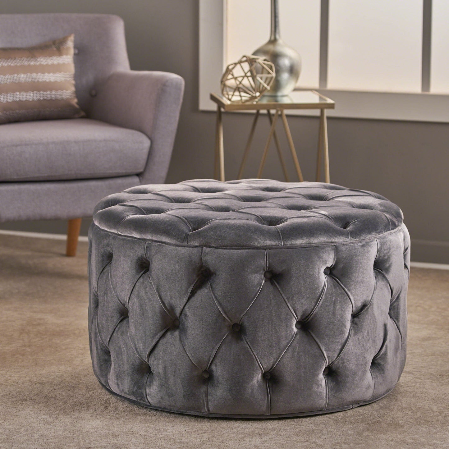 The Modern Glam Ottoman Velvet, featuring a round tufted gray design, rests on a beige carpet in a modern living room near a gray armchair, decorative side table, and a window.