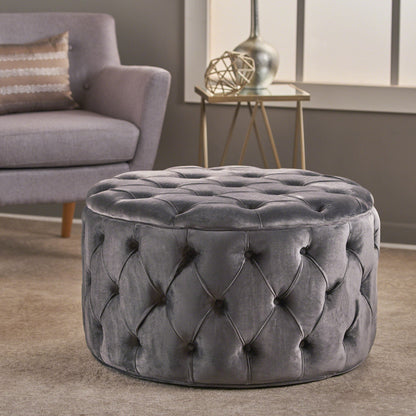 The Modern Glam Ottoman Velvet, featuring a round tufted gray design, rests on a beige carpet in a modern living room near a gray armchair, decorative side table, and a window.