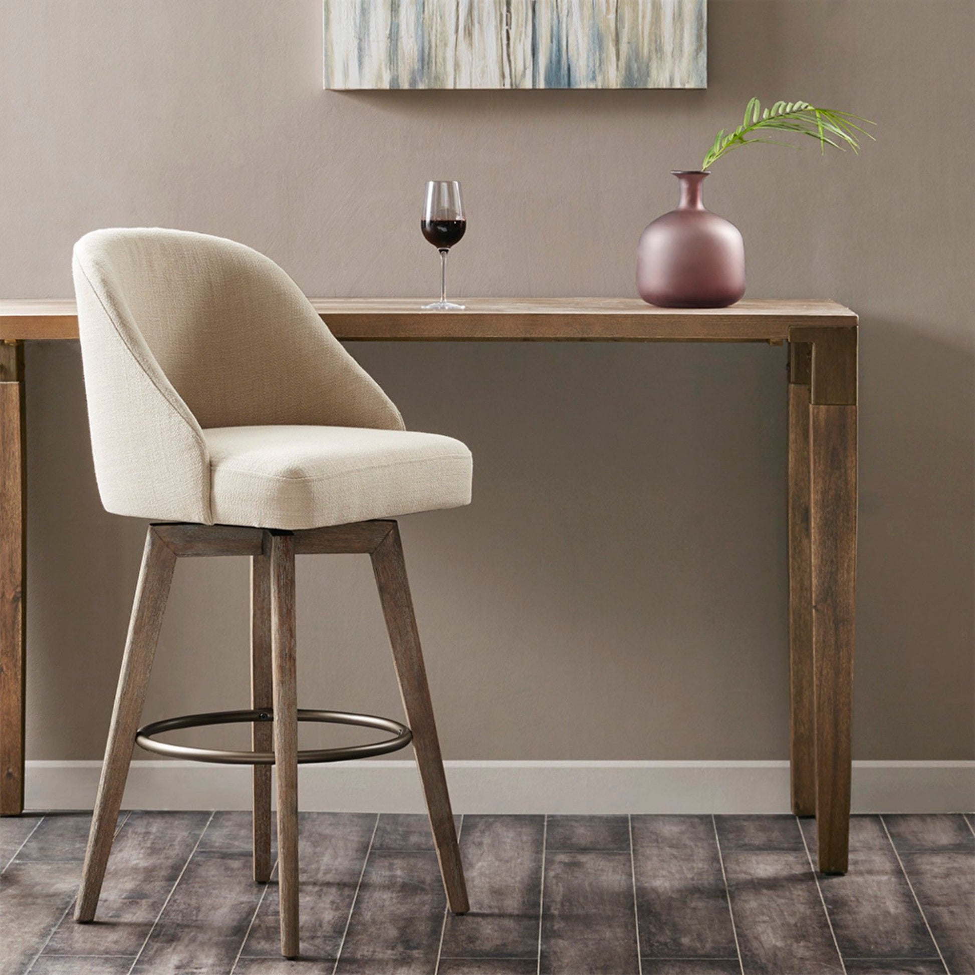 A sand-colored bar stool with a swivel seat sits by a wooden table holding a glass of red wine, a pink vase with a green leaf, and is set against the backdrop of a modern abstract painting on the wall.