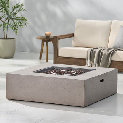 A modern outdoor patio features the Modern 50,000 Btu Square Fire Table With Propane Tank surrounded by lava rocks, a beige cushioned sofa with a gray blanket, a wooden side table, and a potted plant in the background.
