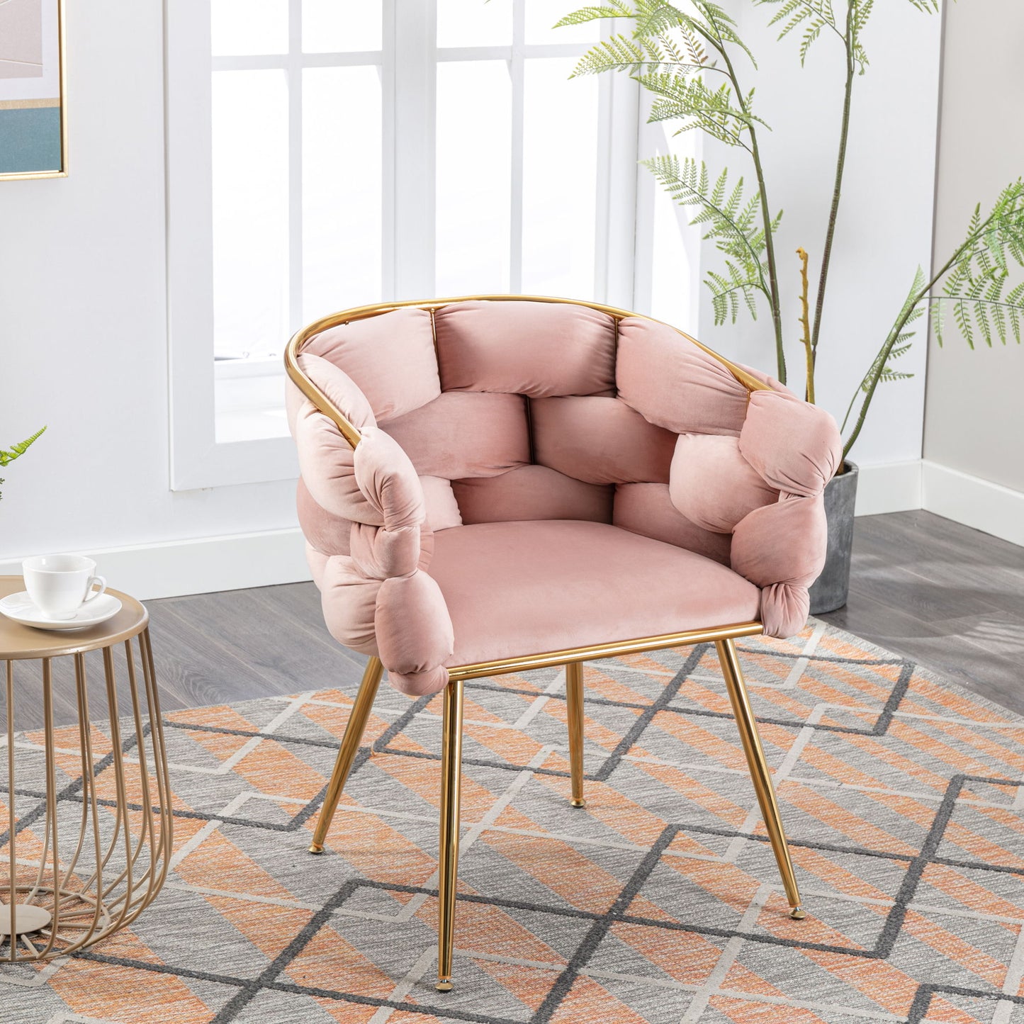 The Luxury Modern Simple Leisure Velvet Single Sofa Chair with pink tufted cushions and gold metal legs is placed on a patterned rug in a bright room with large windows, green plants, and a nearby small round table holding a teacup.