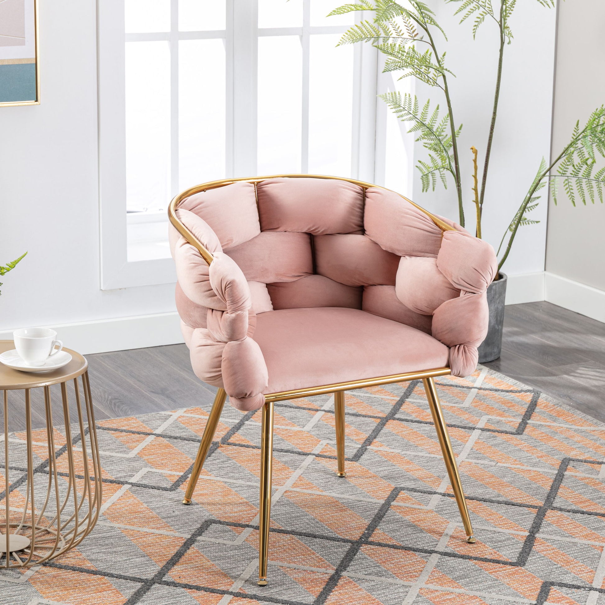 The Luxury Modern Simple Leisure Velvet Single Sofa Chair with pink tufted cushions and gold metal legs is placed on a patterned rug in a bright room with large windows, green plants, and a nearby small round table holding a teacup.