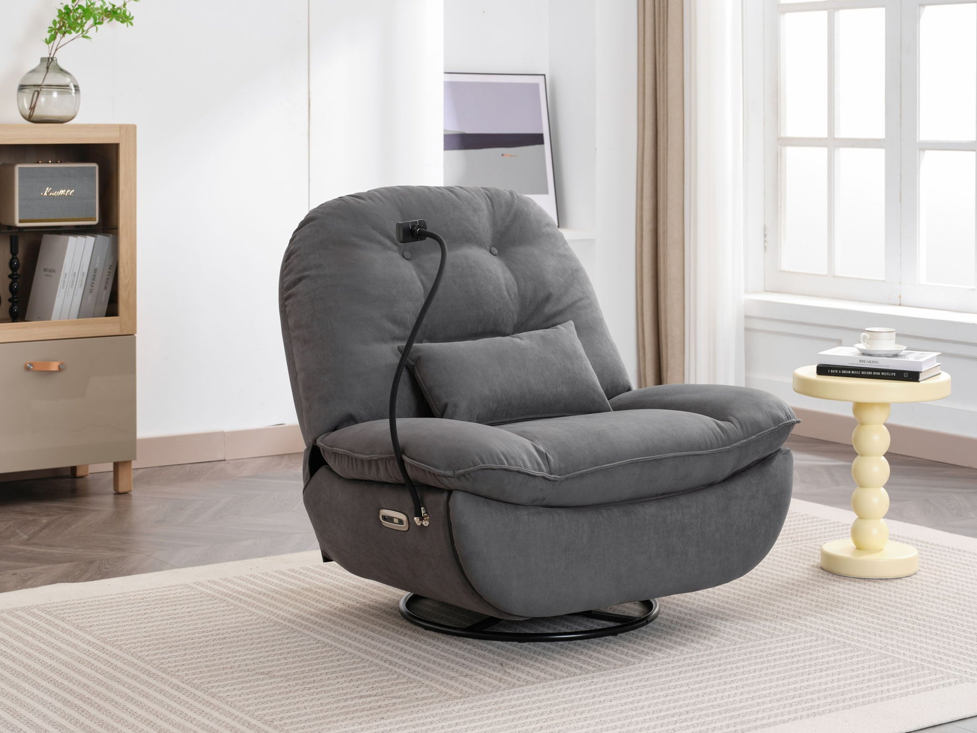 The Power Recliner Swivel Glider USB Charger With Bluetooth Music Player sits on a beige rug in a bright, minimalist living room with a round side table, bookshelf, and large windows.
