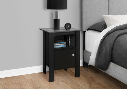 An Accent Nightstand in black with a gray top stands by a bed with gray bedding on a wooden floor, featuring storage, a lamp, clock, books on the shelf, and a lower cabinet for transitional decor.