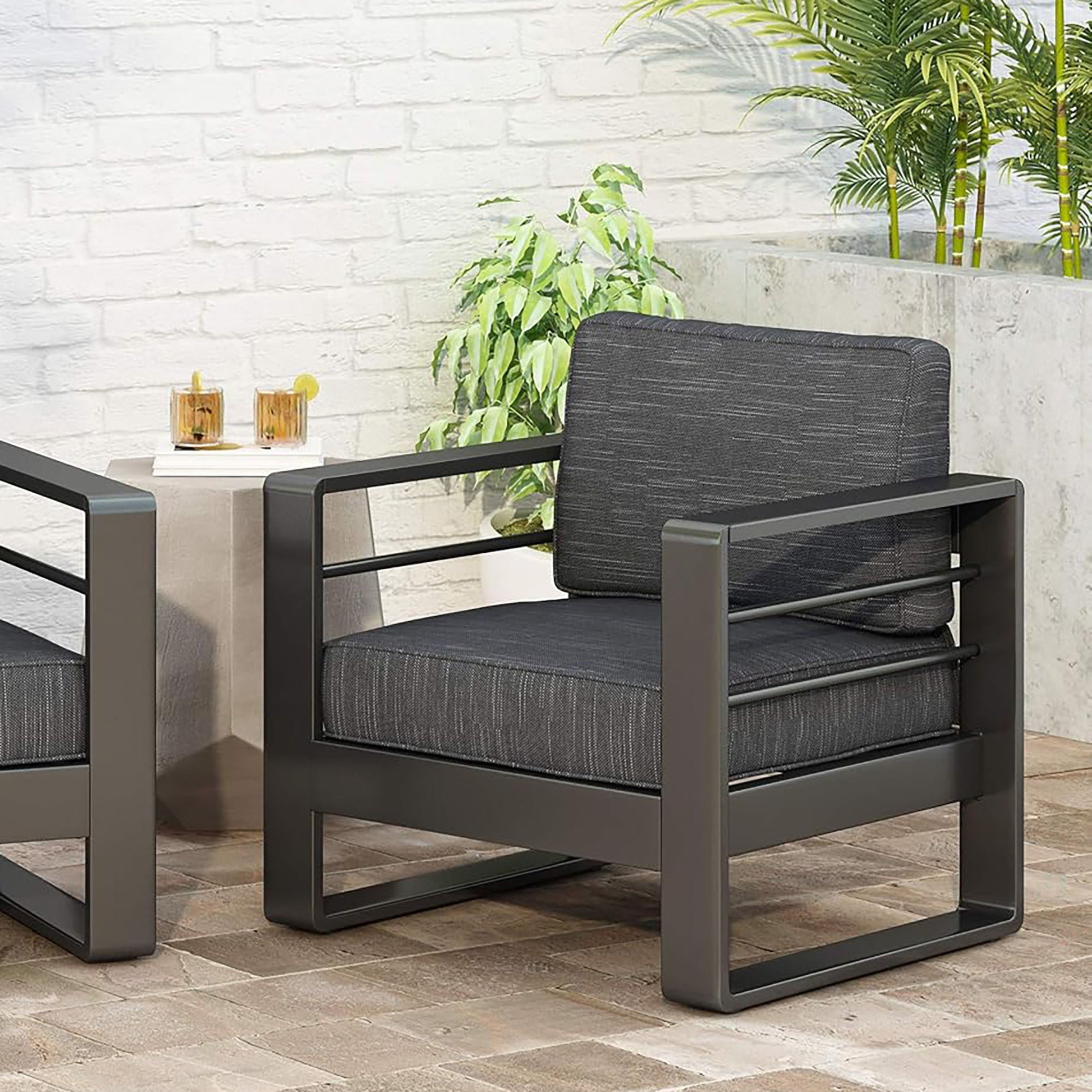 The Maya Bay - Club Chair, featuring dark gray cushions and a metal frame, is placed on a patio next to a small table with two drinks, surrounded by potted green plants and a white brick wall.