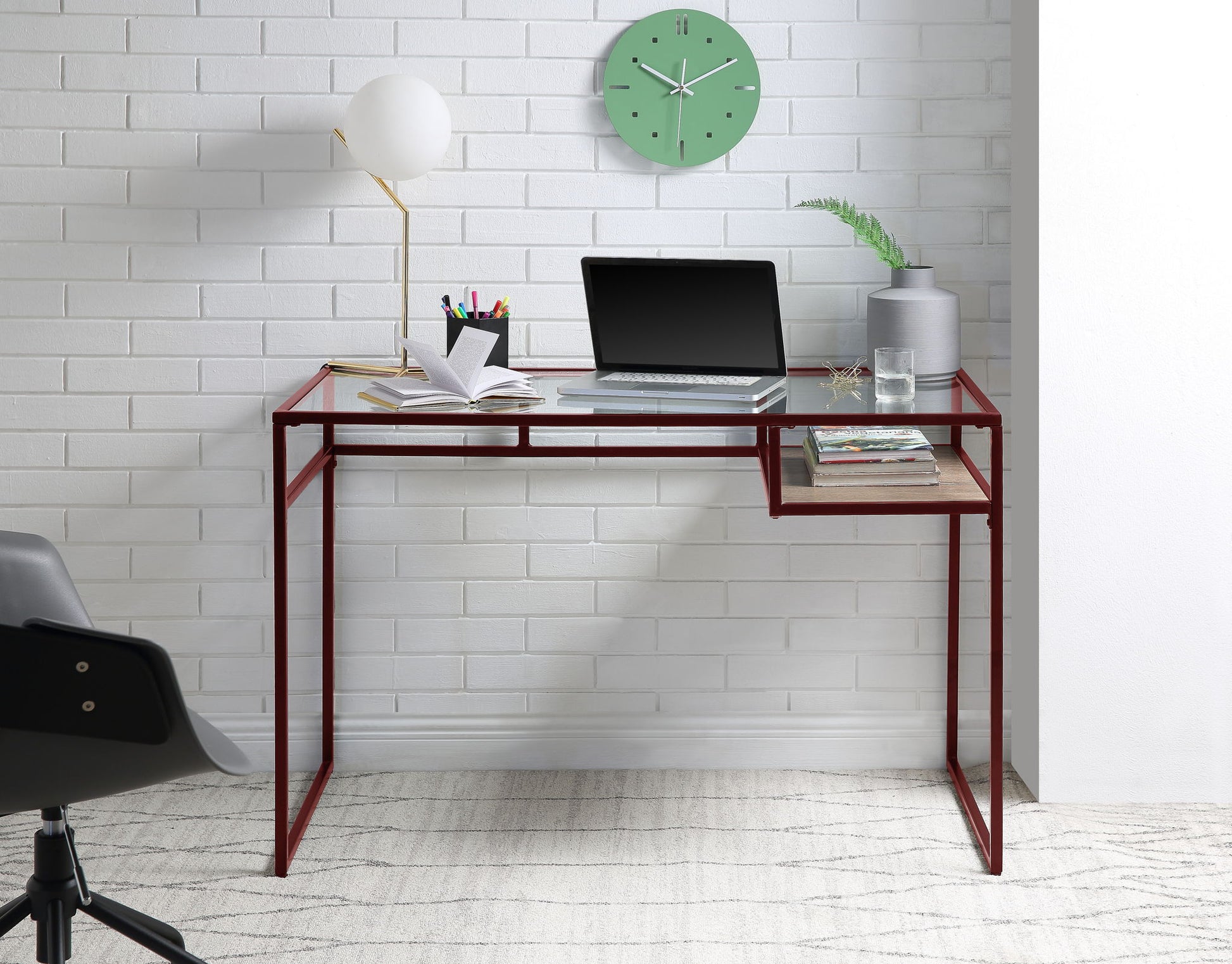 The Yasin - Glass Top Writing Desk is shown with a laptop, books, lamp, plant, and stationery against a white brick wall, a green clock above, and a gray chair partly visible on the left.