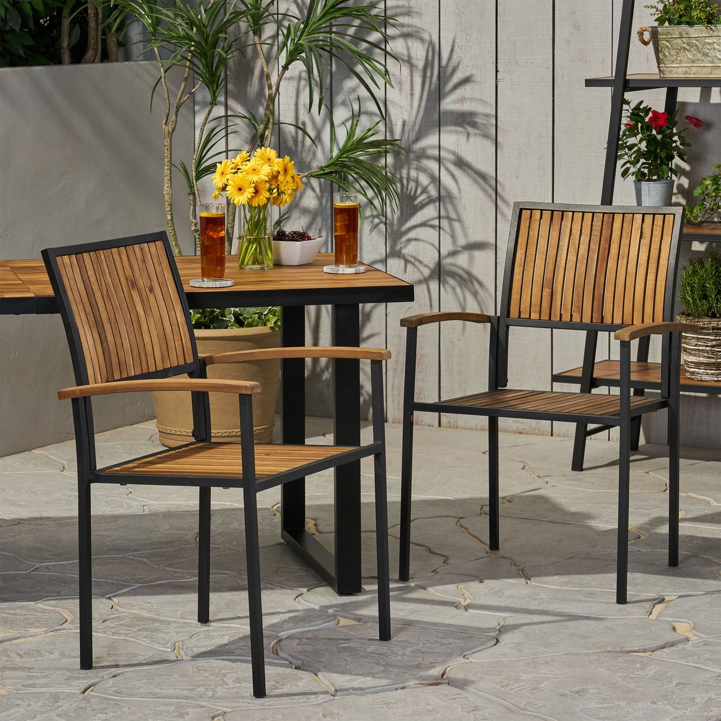Two Vermilion Outdoor Dining Chairs with vertical slats and wooden arms are placed around a patio table set with two glasses of iced tea, a vase of yellow flowers, and a bowl of cherries. Plants and a wooden fence are behind them.