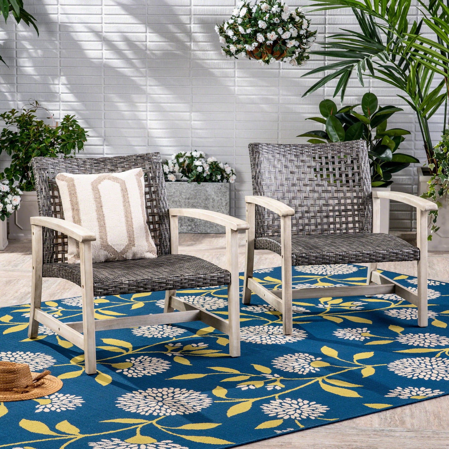 Two gray Outdoor Club Chairs with rattan covers and acacia wood frames rest on a blue floral rug. One features a striped cushion. The cozy patio is brightened by surrounding plants and a white brick wall, with a straw hat on the floor nearby.