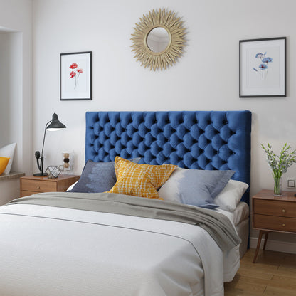 A cozy bedroom features the Annecy Contemporary Upholstered Headboard in navy blue, white bedding, colorful throw pillows, two wooden nightstands, a modern lamp, framed floral art, a sunburst mirror, and fresh flowers on a nightstand.